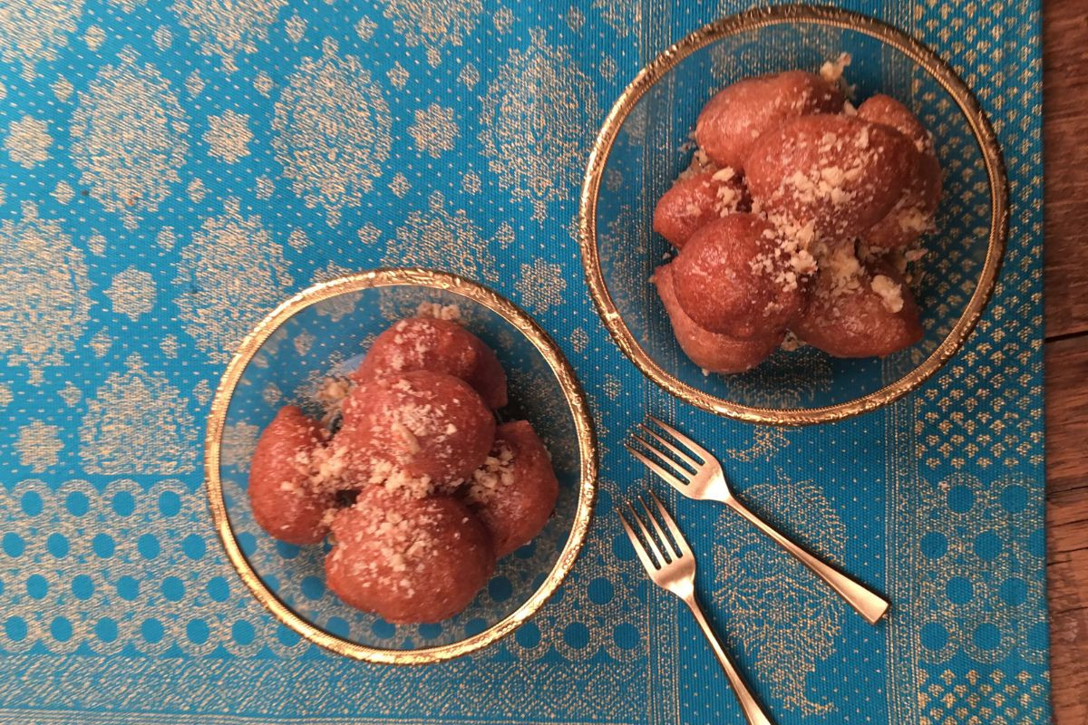 Two bowls of loukoumades, traditional Greek fried dough balls topped with honey and crushed nuts, placed on a vibrant blue patterned tablecloth, with two small forks beside them.